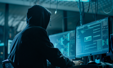 Hooded person hacking into a computer in a dark atmosphere with multiple monitors and cable